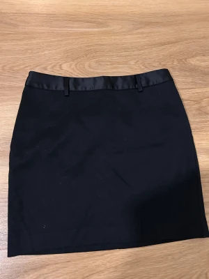 Svart kortkjol från Next, storlek 36 - Svart kortkjol från Next i storlek 36. Kjolen har en stilren design med slät yta och ett glansigt band upptill med hällor för bälte. Materialet känns mjukt och har en lätt struktur, perfekt för dig som gillar minimalistisk stil.