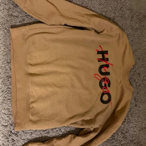 Beige sweatshirt från Hugo med logga - Snygg beige sweatshirt från Hugo med stor svart och röd logga framtill. Tröjan har rund hals, ribbade muddar vid ärmslut och nederkant samt lång ärm. Tillverkad i mjuk bomull som är skön mot huden.  Storlek M  och kostar runt 2300kr knappt använd