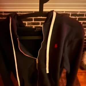 Mörkblå hoodie från Polo Ralph Lauren - Mörkblå hoodie från Polo Ralph Lauren med vit snörning och dragkedja framtill. Klassisk röd logga broderad på bröstet. Tröjan har huva och är långärmad, perfekt för chill dagar. Mjuk bomullskänsla och stilren design.