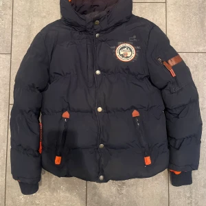 Mörkblå pufferjacka Geographical Norway - Snygg mörkblå pufferjacka från Geographical Norway med orange detaljer och broderad patch på bröstet. Jackan har hög krage, dragkedja och knappar framtill, samt fickor med dragkedja och ribbade muddar vid ärmslut. Perfekt för kalla dagar. Dock är den sluten under armhålan på högerarm. Kom gärna med prisförslag😊