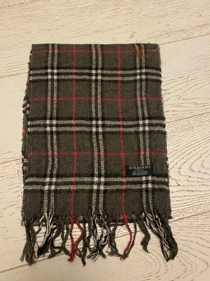 Rutig halsduk från Burberry i kashmir - Snygg halsduk från Burberry i 100% kashmir, tillverkad i England. Den har ett klassiskt rutigt mönster i mörkgrått, vitt och rött med fransar i ändarna. Perfekt för att ge din outfit en lyxig och tidlös touch.