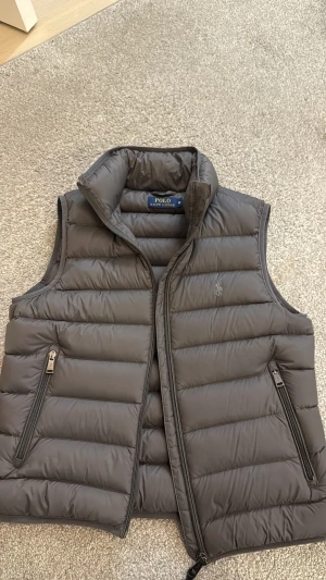 Grå dunväst från Polo Ralph Lauren - Snygg grå dunväst från Polo Ralph Lauren med quiltad design och hög krage. Västen har två praktiska fickor med dragkedja och diskret logga på bröstet. Perfekt att slänga över en hoodie eller långärmad tröja för extra värme. Nypris 3499. Säljs för 1000