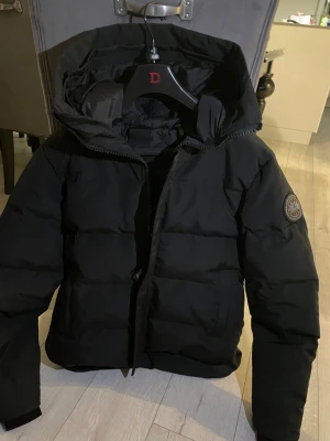 Canada Goose Black Label - Säljer en svart dunjacka från Canada Goose med huva och tydlig logotyp på ärmen. Jackan har dragkedja, knappar och två stora fickor framtill. Den är quiltad och har en clean look som är perfekt för kalla dagar. Det är den nya black label modellen. Den är i princip aldrig använd utan bara testad. Skriv vid intresse och vid önskan av fler bilder! Pris går att diskutera.
