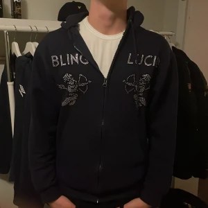 Mörkblå Bling Lucid hoodie med strass - Mörkblå hoodie från Bling Lucid i storlek S, med strassdetaljer och motiv av änglar på bröstet. Tröjan har huva, dragkedja och är tillverkad i mjuk bomull och polyester. Perfekt för dig som gillar lite extra bling och streetstyle.