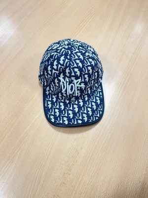  Dior keps med monogram - Säljer en snygg keps från Dior med broderad Dior-logga framtill. Kepsen har böjd skärm och justerbar rem baktill. Tillverkad i bomullstyg med exklusiv känsla, perfekt för dig som vill sticka ut med en ikonisk accessoar. Hel oanvänd