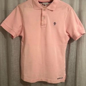 Rosa Moncler pikétröja i bomull - Snygg ljusrosa pikétröja från Moncler i storlek S. Tröjan har klassisk krage, korta ärmar och knappar framtill. Moncler-logga broderad på bröstet och nedtill. Tillverkad i mjuk bomull som är skön mot huden. Perfekt för dig som gillar stilrena och trendiga plagg.