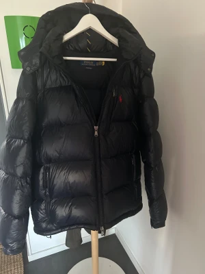 Svart pufferjacka Polo Ralph Lauren XL - Svart pufferjacka från Polo Ralph Lauren i storlek XL. Jackan har en hög krage, huva, två sidofickor med dragkedja och klassisk röd Polo-logga på bröstet. Typ helt oanvänd använd cirka 2 ggr, ska sälja då han köpt en ny jacka. Brukar ha L i storlek men XL passar perfekt.