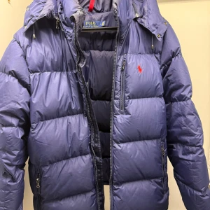 Ralph Lauren puffer jacket  - Säljer en blå dunjacka från Ralph Lauren.   Jackan har huva, flera dragkedjefickor och är perfekt för den kommande vintern. Passformen är normal och storlek M sitter bra på mig som är 185. Jackan har används sparsamt men tyvärr har den en defekt på ena ärmen där det gått hål. Detta är lagat men syns, se sista bilden. Hör av dig vid fler frågor! Pris kan diskuteras