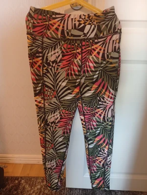 DKNY Sport leggings Army Palm S - Snygga leggings från DKNY Sport i storlek S med tropiskt palmbladsmönster i grönt, rosa, gult och svart. Modellen är tight och högmidjad, perfekt för träning. Materialet är mjukt och stretchigt syntet, vilket ger en skön passform och rörelsefrihet.
