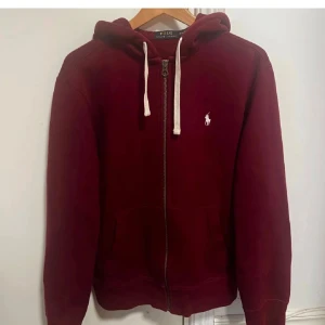 Vinröd hoodie från Polo Ralph Lauren - Vinröd hoodie från Polo Ralph Lauren med dragkedja och vit broderad logga på bröstet. Tröjan har huva med vita snören och två fickor framtill. Perfekt för en chill och stilren look.