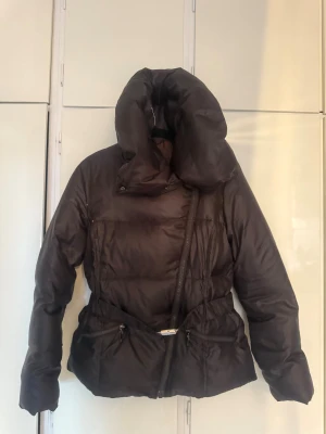 Brun pufferjacka med stor krage - Säljer en svart pufferjacka med riktigt fet och hög krage som ger extra mysfaktor. Jackan har dragkedja i sned design, två fickor med dragkedja och ett bälte i midjan med metallspänne. Materialet är glansigt och känns syntetiskt, perfekt för kalla dagar.