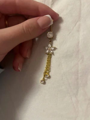 Guldig belly piercing - Säljer en lång belly piercing i guld med glittrande vita stenar och en blomformad detalj. Hängande kedjor med små pärlor ger extra bling och rörelse.