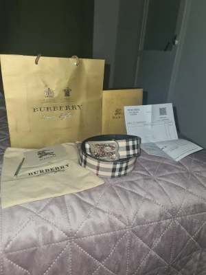 Burberry rutig skinnbälte med B-spänne - Säljer ett snyggt Burberry bälte i klassiskt rutigt mönster med beige, svart och röd färg. Bältet har ett exklusivt B-spänne i silver och guld med Burberrys ikoniska ryttare. Materialet är skinn och bältet kommer med originalpåse, box och certifikat.