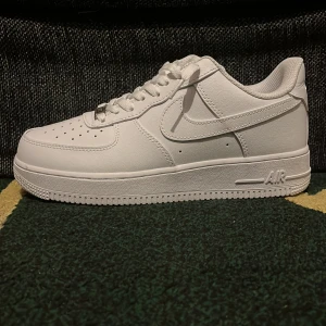 Nike Air Force 1 helvita sneakers - Klassiska Nike Air Force 1 sneakers i helvitt skinn med vit sula och snörning. Ikonisk siluett med perforerad tå och Nike Swoosh på sidorna. Perfekta för dig som vill ha en clean och tidlös look. Passar till allt och är superbekväma.