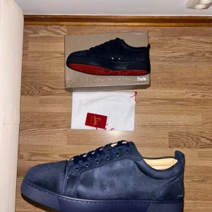 Christian Louboutin svarta sneakers mocka - Säljer ett par svarta sneakers från Christian Louboutin i mjuk mocka med klassisk röd sula och rund tå. Skorna har snörning och diskret broderad logga på hälen. Insidan är beige och de har en platt sula. Perfekt för dig som gillar lyxiga detaljer.
