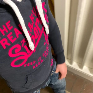 Superdry Hoodie (sällsynt) - Snygg svart hoodie från Superdry med stort rosa tryck på bröstet och vita snören i huvan. Tröjan har känguruficka framtill och ribbade muddar vid ärmslut och nederkant. Den har inga defekter och är som ny. Om du undrar något är det bara att fråga🙌✅