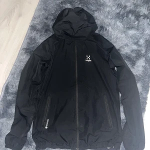 Svart Haglöfs Gore-Tex vindjacka - Svart vindjacka från Haglöfs med huva och hel dragkedja framtill. Jackan är tillverkad i Gore-Tex-material som är både vatten- och vindtätt, perfekt för blåsiga och regniga dagar. Två sidofickor med dragkedja och meshfoder på insidan för extra komfort.