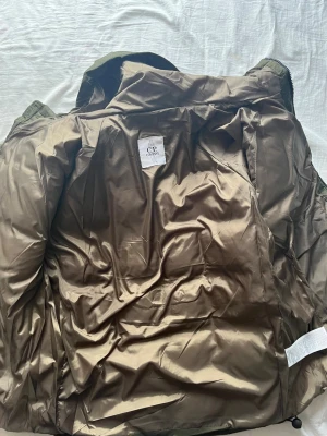 Olivgrön pufferjacka från C.P. Company - Säljer en olivgrön pufferjacka från C.P. Company med huva och dragkedja. Jackan har quiltad design och en ikonisk linsdetalj på ärmen. Materialet är syntet och polyester, perfekt för kalla dagar. Snygg och funktionell med justerbar huva och praktiska fickor.