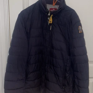 Mörkblå lättviktsdunjacka Parajumpers XL - Mörkblå superlätt dunjacka från Parajumpers i storlek XL. Jackan har quiltad design, hög krage, dragkedja framtill och ikonisk gul detalj vid halsen. Märkespatch på ärmen och två dragkedjefickor. Perfekt för kyliga dagar. Nackdel är att högerfickan är sönder, alltså kan du inte placera några föremål. Vänster funkar perfekt. Annars är skicket bra på jackan.
