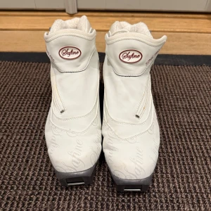 Vita Safine Sonic Pro pjäxor TecnoPro - Vita Safine Sonic Pro pjäxor från TecnoPro med grå detaljer och dragkedja på sidan. Fodrade med mjukt, vitt material för extra värme. Snyggt broderad logga och robust sula med SNS Profil-system. Perfekta för längdskidåkning under vintern.