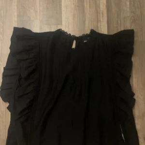 Svart blus med volang från Gina Tricot - Svart blus från Gina Tricot i storlek M med snygga volangdetaljer längs axlarna och ärmarna. Blusen har en rund hals med spetskant och en liten knapp i nacken. Luftigt matrial är knappt använd.💗💗