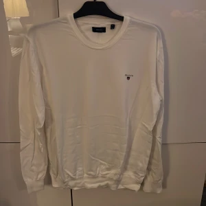 Vit långärmad tröja från GANT XL - Vit långärmad tröja från GANT i storlek XL. Tröjan har rund halsringning och ett litet broderat GANT-logo på bröstet. Mjuk och skön känsla, perfekt för chill dagar. Ribbstickade muddar vid ärmslut och nederkant ger en snygg passform.