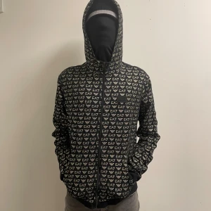 Svart EA7 vindjacka med logomönster - Svart vindjacka från EA7 Emporio Armani med hel dragkedja och huva. Jackan har ett all-over mönster med EA7-loggor.