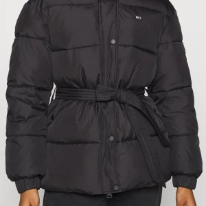 Svart pufferjacka från Tommy Jeans - Svart pufferjacka från Tommy Hilfiger med huva och bälte i midjan. Jackan har quiltad design, tryckknappar framtill och ett litet logomärke på bröstet. Perfekt för kalla dagar med sin tjocka och fluffiga look. Långärmad och med elastiska muddar vid ärmslut.