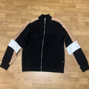 Svart bomberjacka från BOSS - Snygg svart bomberjacka från BOSS med beige och vita paneler på ärmarna. Jackan har hel dragkedja framtill, ribbade muddar och två fickor. Materialet är mjukt och känns som polyester, perfekt för en chill och stilren look.KÖP HELA DRESSEN OCH FÅ DEM FÖR 3000kr