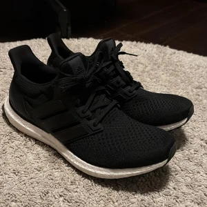 Adidas Ultraboost svarta sneakers - Svarta Adidas Ultraboost sneakers med stickad ovandel och klassiska tre ränder på sidan. Skorna har snörning, vit Boost-mellansula och en svart yttersula med grepp. Perfekta för dig som gillar sportig stil och komfort. Materialet är syntet och textil.