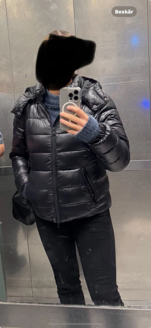 Svart pufferjacka med huva från lager 157 - Säljer min svarta pufferjacka från lager 157 med huva och dragkedja framtill. Jackan har ett quiltat mönster och är tillverkad i glansig polyester. Perfekt för kalla dagar och har en snygg, normal passform. Fickor med dragkedja på sidorna för extra funktion.
