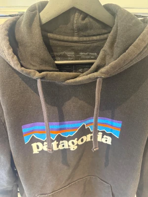 Svart Patagonia hoodie med tryck - Svart hoodie från Patagonia med klassiskt färgglatt logotryck på bröstet i bra skick. Köpt från Zalando, skriv för fler bilder. 