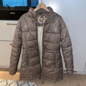 Brun pufferjacka med dragkedja - Säljer en brun pufferjacka med hög krage och långa ärmar. Jackan har två stora fickor med dragkedja framtill och quiltad design. Perfekt för kyliga dagar och har en snygg, enkel look.