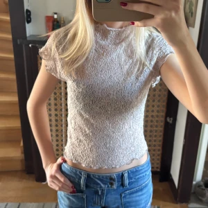 Ljusbeige spetsig blus med volang - Supersöt ljusbeige blus med spetsmönster och volangkant vid ärmar och nederkant. Croppad passform och korta ärmar, perfekt att styla med jeans eller kjol. Hög hals och genombrutet tyg ger en romantisk vibe.