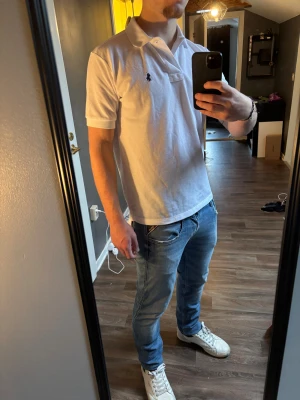 Vit pikétröja från Ralph Lauren - Snygg vit pikétröja från Ralph Lauren med klassisk krage och korta ärmar. Tröjan har den ikoniska broderade loggan i svart på bröstet.