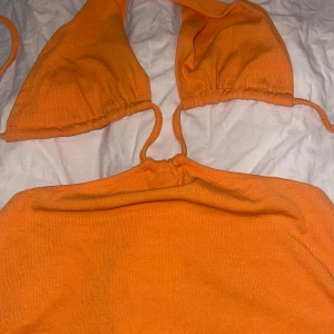 Orange klänning med och snörning - Snygg orange klänning med omlott-topp och knyt i nacken. Klänningen har en tight passform och är tillverkad i ett stretchigt syntetmaterial. Perfekt för stranden eller poolen, med trendig och ungdomlig look.