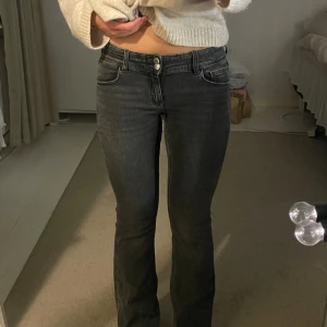 Gråa bootcut jeans med fickdetaljer - Snygga svarta jeans med bootcut passform och låg midja. Jeansen har klassiska fem fickor, varav bakfickorna har lock och knappdetalj. Materialet är jeans med en lätt tvättad look och silverfärgade knappar framtill.