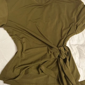 Olivgrön topp med knyt i midjan - Snygg olivgrön topp med korta ärmar och rund hals. Toppen har en cool knytning i midjan som ger en extra detalj och form till plagget. Materialet känns mjukt och lätt, perfekt för dig som gillar enkel men trendig stil.