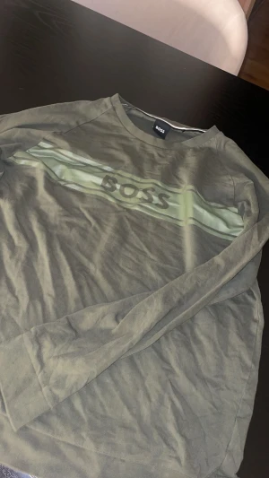 Grön långärmad tröja från BOSS - Snygg grön långärmad tröja från BOSS med ett stilrent tryck över bröstet i form av en ljusgrön rand och loggan. Tröjan har rund hals och är gjord i mjuk bomull, perfekt för chill dagar. Passar dig som gillar enkel och clean stil.