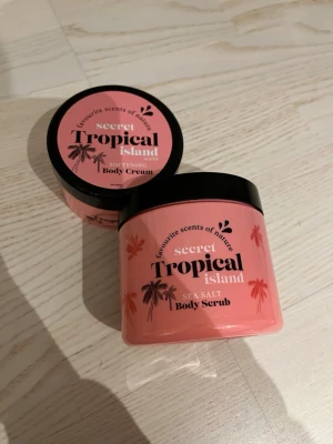 Secret Tropical Island bodykit - Säljer ett set med body scrub och body cream från Favourite Scents of Nature i doften Secret Tropical Island. Båda burkarna är rosa med svarta lock och har palmmotiv. Body scrub innehåller havssalt och båda produkterna ger en tropisk känsla.