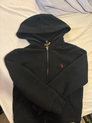 Svart hoodie från Polo Ralph Lauren - Klassisk svart hoodie från Polo Ralph Lauren med dragkedja framtill och huva. Den har det ikoniska röda Polo-logot på bröstet och två fickor fram. Tillverkad i mjukt bomullsmaterial som är skönt att ha på sig. Barnstorlek men passar mig som vanligtvis har xs