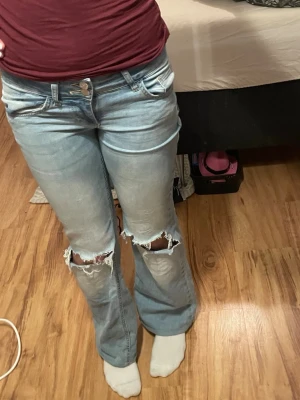 Ljusblå bootcut jeans med slitningar - Säljer ett par ljusblå bootcut jeans med slitna hål över knäna och klassiska fem fickor. Jeansen har låg midja och är tillverkade i bomull med en skön jeanskänsla. Perfekta för en avslappnad och trendig look. Dom är uppsydda så dom är lite kortare för att passa bättre, passar mig perfekt och jag är 160 cm🙌🏼 Dom har även fickor med knappar där bak! 
