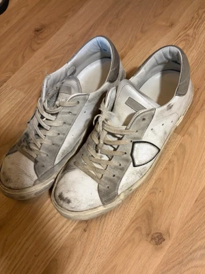 Vita och grå sneakers från Philippe Model - Säljer ett par klassiska Philippe Model sneakers i vitt och grått skinn med den ikoniska sköld-loggan på sidan. Skorna har snörning, rund tå och platt sula. Perfekta för dig som gillar stilrena och tidlösa sneakers med en sportig touch.