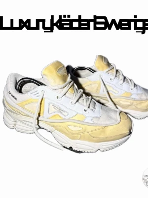 Raf Simons x Adidas Ozweego sneakers - Säljer ett par Raf Simons x Adidas Ozweego sneakers i vitt och ljusgult med cool futuristisk design. Skorna har snörning och detaljerad sula, samt branding på sidan. Perfekta för dig som gillar statement sneakers och streetwear.