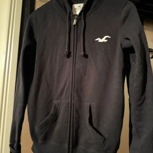 Svart hoodie från Hollister - Svart zip-hoodie från Hollister med vit text 