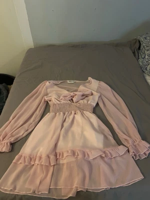 Ljusrosa volangklänning Designby Amor - (Mer Rosa i verkligheten) Supersöt ljusrosa kortklänning från Designby Amor med långa transparenta ärmar, volangdetaljer och smock i midjan. Klänningen har v-ringning med knyt framtill. Använd 1gång på min avslutning🩷🩷nypris 500kr