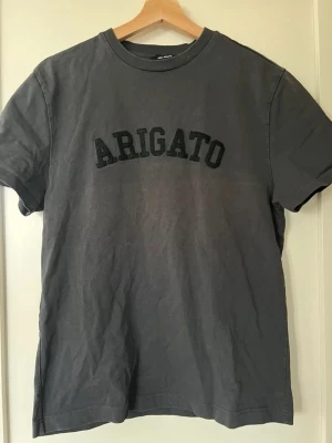 Grå t-shirt från Axel Arigato - Snygg grå t-shirt från Axel Arigato med svart text på bröstet. Klassisk rund halsringning och korta ärmar. Skön enkel tröja med lite användning.OBS: STOR I STORLEKEN. Först till kvarn!