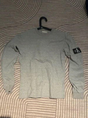 Grå långärmad tröja från Calvin Klein - Snygg grå långärmad tröja från Calvin Klein med rund halsringning och en svart CK-logga på vänster ärm. Tillverkad i mjukt bomullsmaterial som känns skönt mot huden. Enkel och stilren design som passar till det mesta.