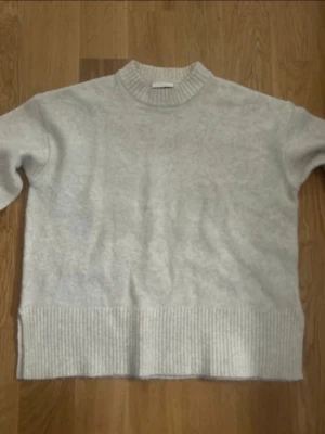 Ljusgrå stickad tröja med ribbade detaljer - Mysig ljusgrå stickad tröja med rund halsringning och breda ribbade muddar vid ärmslut och nederkant. Tröjan har en avslappnad passform och är perfekt för kyliga dagar. Materialet känns mjukt och behagligt mot huden.