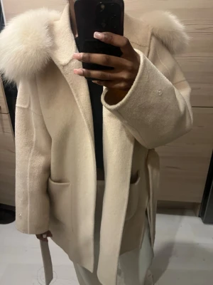 Beige kappa med äkta päls - Stilren beige kappa med stora fickor, knytskärp i midjan och fluffiga fuskpälsdetaljer på huvan. Kappan har en loose passform och är perfekt för kyligare dagar. Materialet är mjukt och ser ut att vara ullblandning.